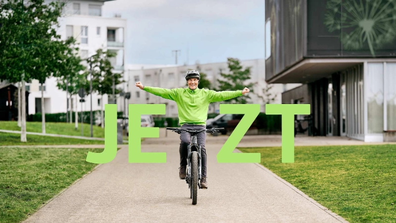 Radler fährt strahlend auf seinem Fahrrad mit gestreckten Armen mit Schriftzug "Jetzt"