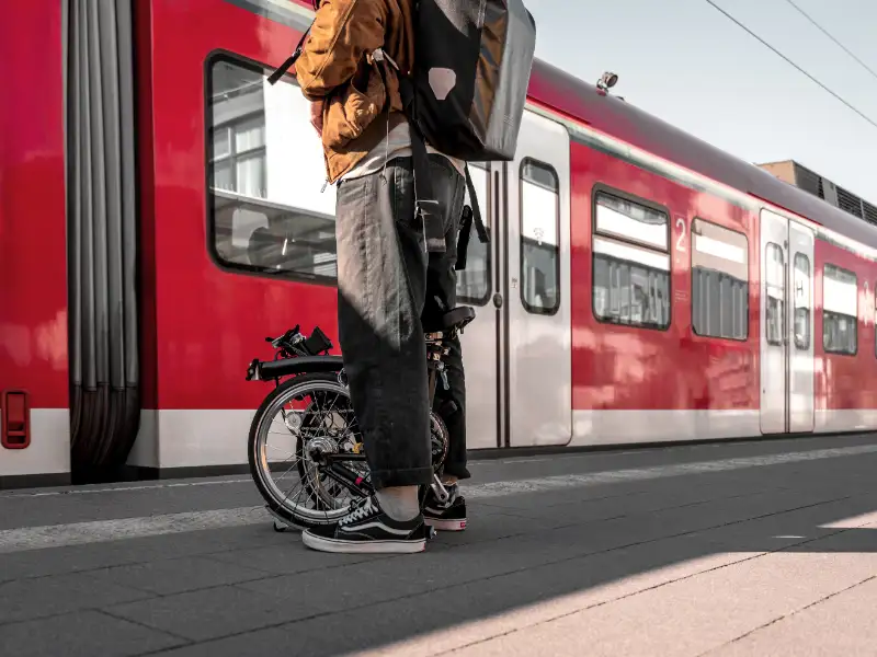 Ein JobRadler steht mit einem Faltrad der Marke Brompton auf dem Bahnsteig vor einem Inter-City