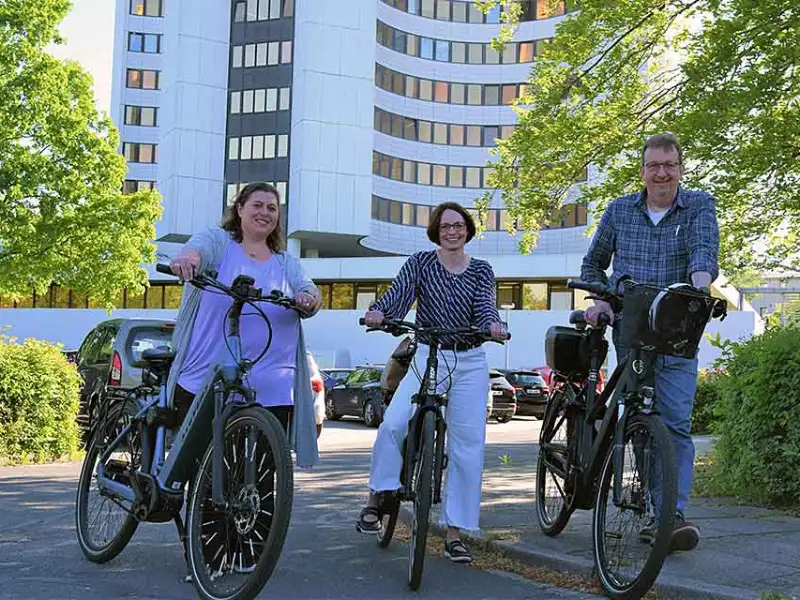 Drei Mitarbeiter:innen, die an einem sonnigen Tag mit dem Fahrrad in Dithmarschen vor einem großen Gebäude stehen Drei Mitarbeiter:innen, die an einem sonnigen Tag mit dem Fahrrad in Dithmarschen vor einem großen Gebäude stehen