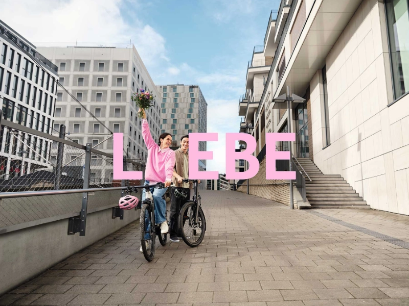 2 JobRadler:innen auf einem Fahrrad in einem urbanen Umfeld mit einem Blumenstrauß.