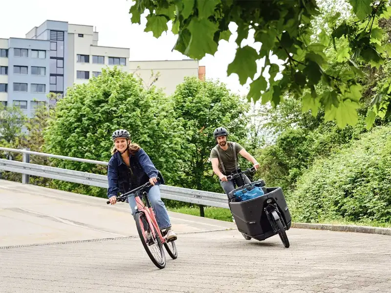 JobRadlerin und JobRadler fahren fröhlich um eine Kurve mit Citybike und Lastenrad JobRadlerin und JobRadler fahren fröhlich um eine Kurve mit Citybike und Lastenrad