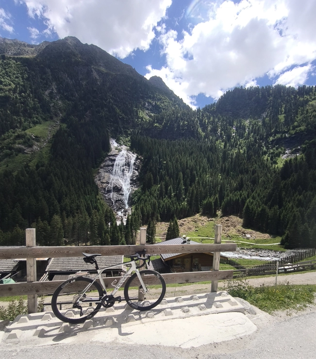 Weißes Rennrad an einem Holzzaun mit Blick auf einen Wasserfall, grüne Berglandschaft und bewaldete Hänge unter blauem Himmel.