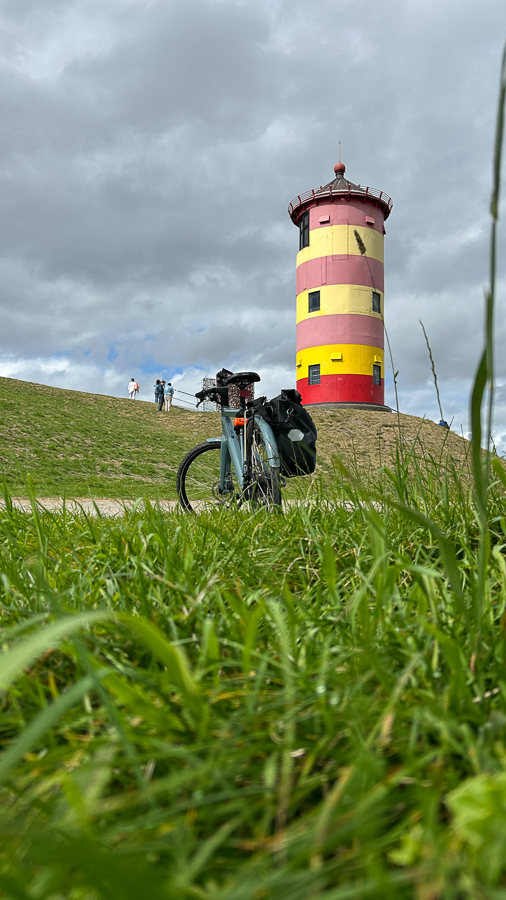 Ein bepacktes Fahrrad steht auf einer grünen Wiese vor einem rot-gelb gestreiften Leuchtturm unter bewölktem Himmel.