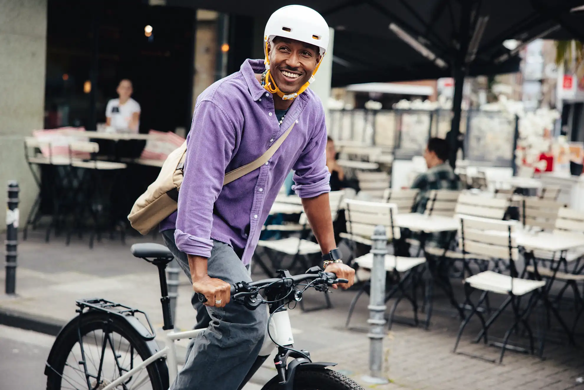 Mann fährt lachend in urbaner Umgebung mit seinem Rad, im Hintergrund ein Cafe