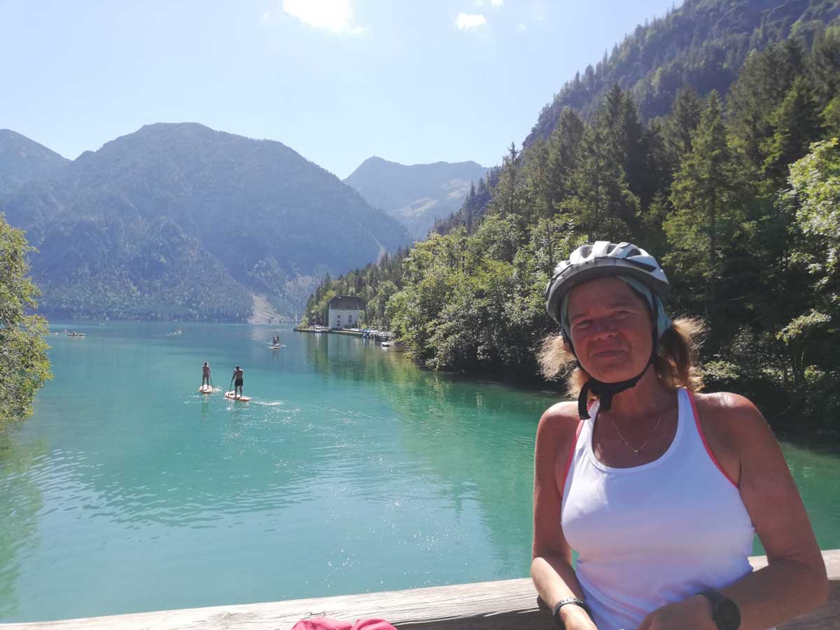 JobRad Moment von Brigitte mit Helm und weißem Top, im Hintergrund zwei Paddler auf dem See und Berge