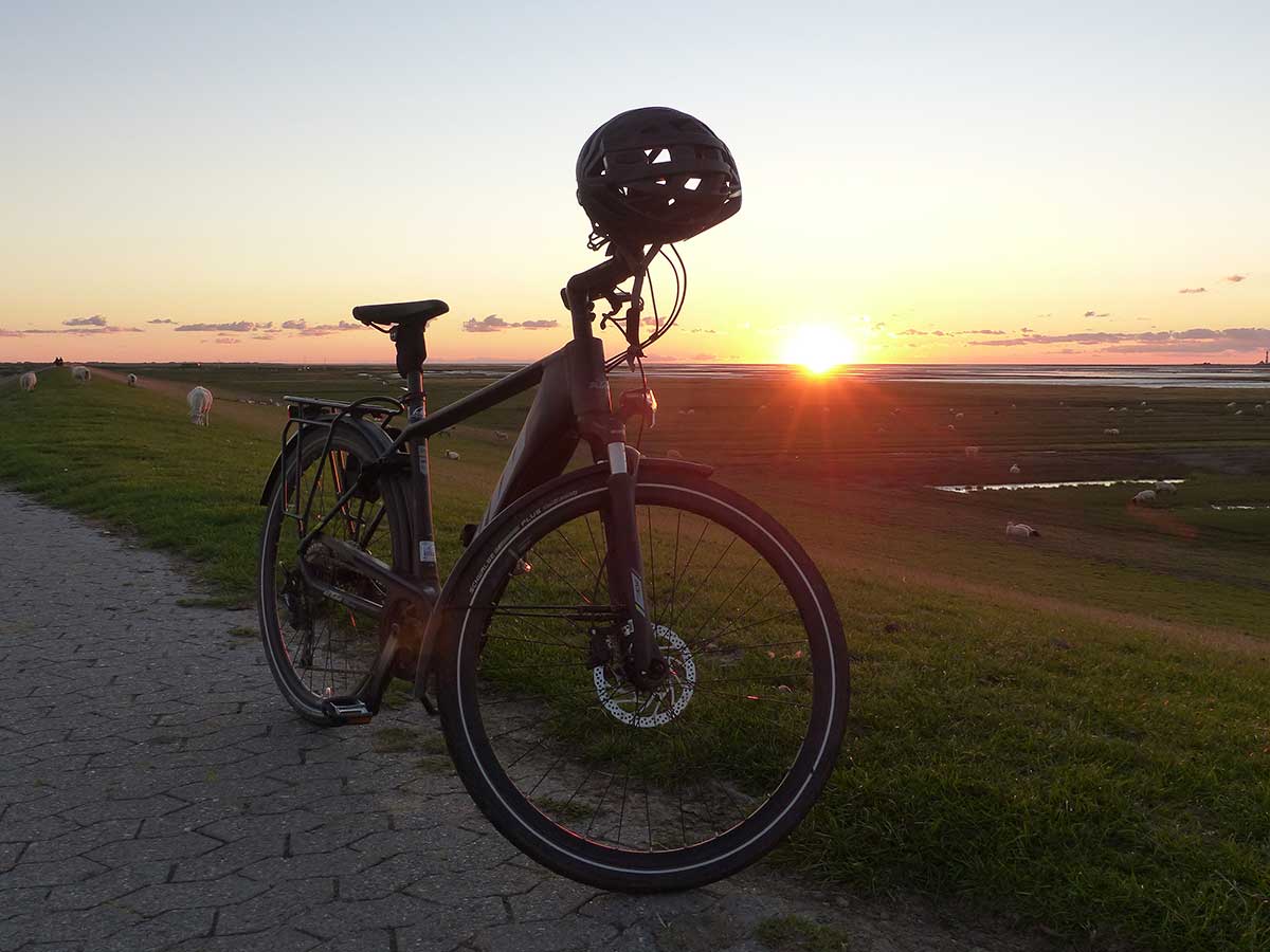 JobRad Moment von Thomas mit E-bike it Helm am Lenker auf gepflastertem Weg, im Hintergrund geht die Sonne in der Ebene unter,